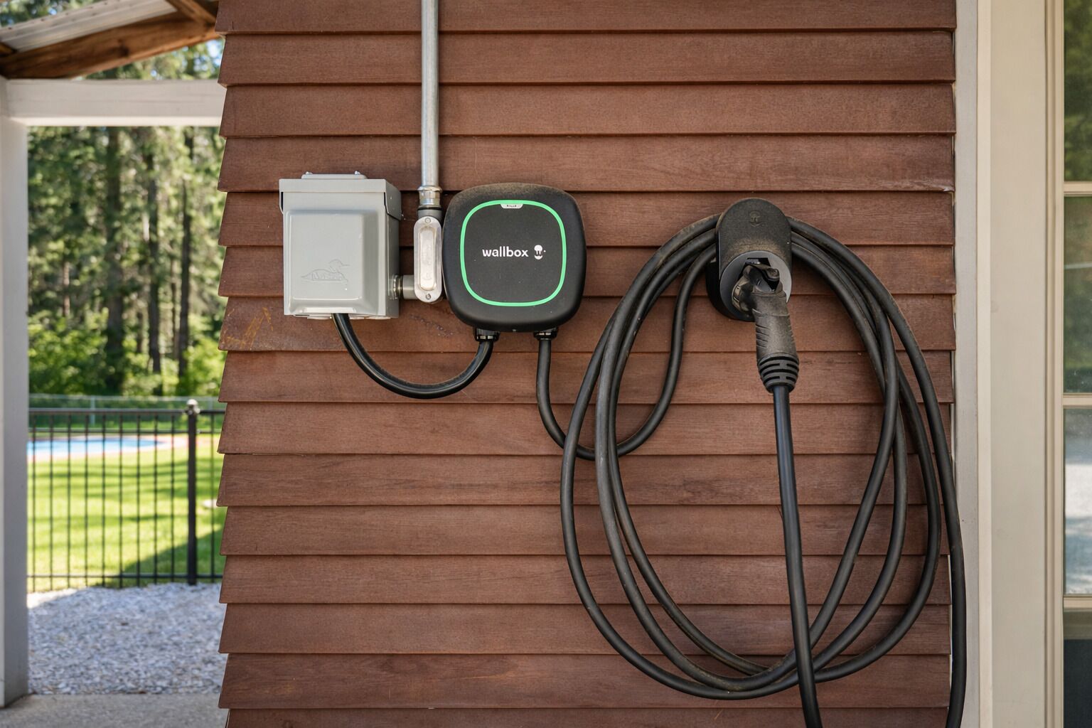 Een laadstation voor elektrische voertuigen bevestigd aan een houten muur, met een metalen leiding en een kabel aangesloten op een wandcontactdoos.