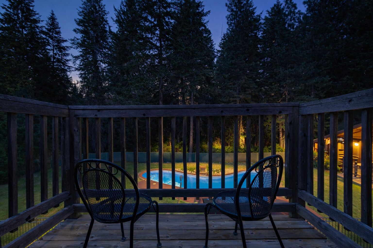 A wooden deck with two chairs facing a swimming pool, surrounded by trees.