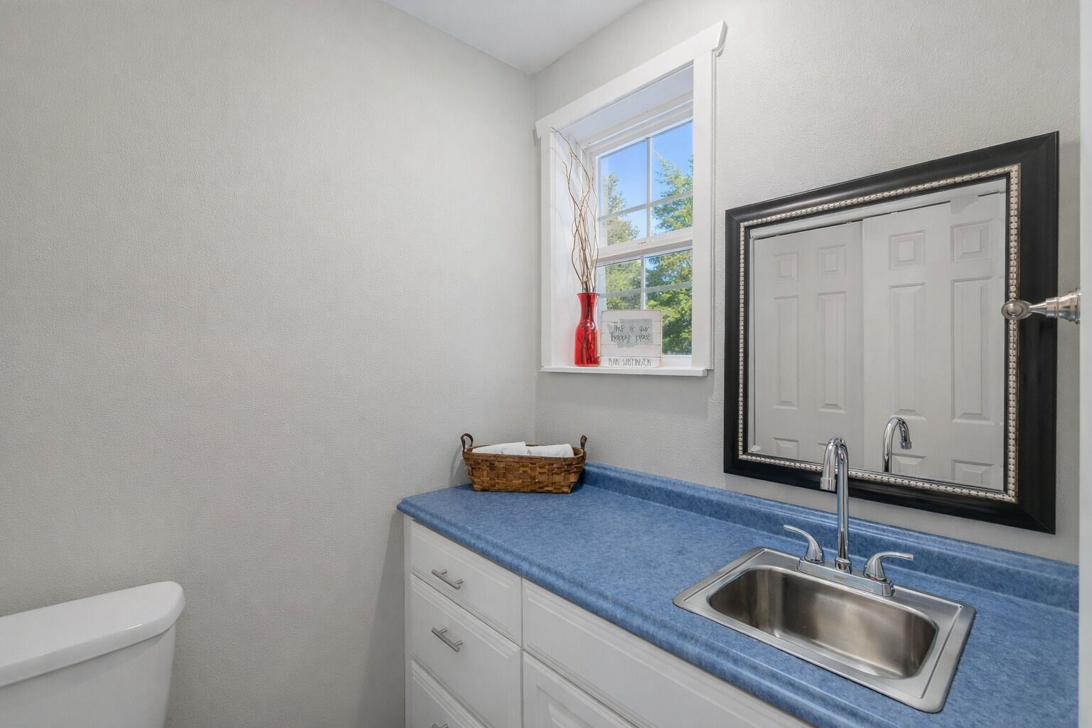 A bathroom with a white toilet, a blue countertop, a stainless steel sink, a mirror with a decorative frame, and a window with a view of trees.