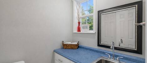 A bathroom with a white toilet, a blue countertop, a stainless steel sink, a mirror with a decorative frame, and a window with a view of trees.