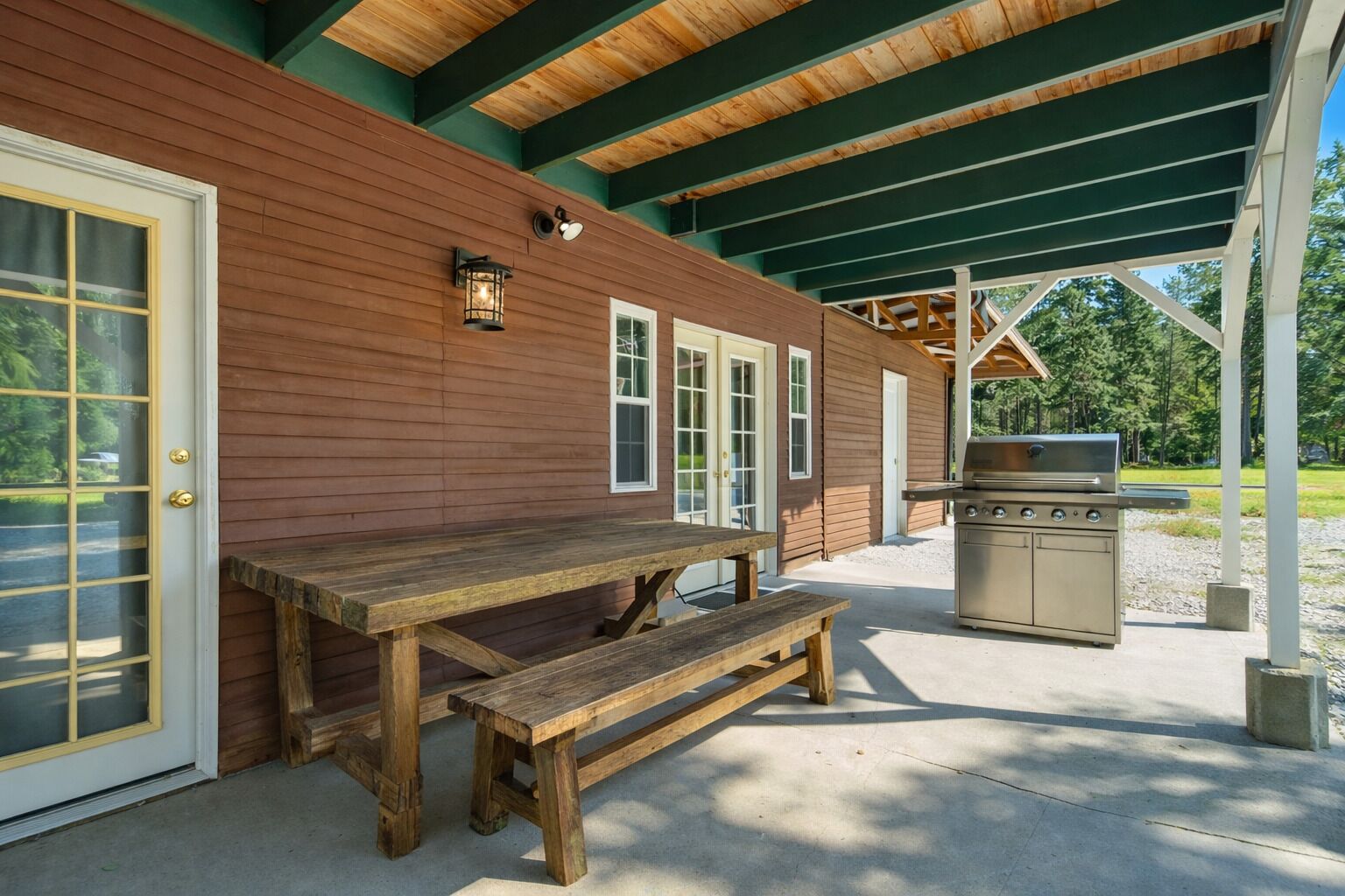 A covered outdoor area with a wooden table and bench, a grill, and a building with a green roof and wooden siding.
