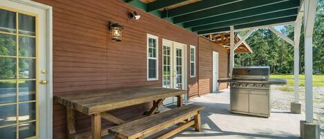 A covered outdoor area with a wooden table and bench, a grill, and a building with a green roof and wooden siding.