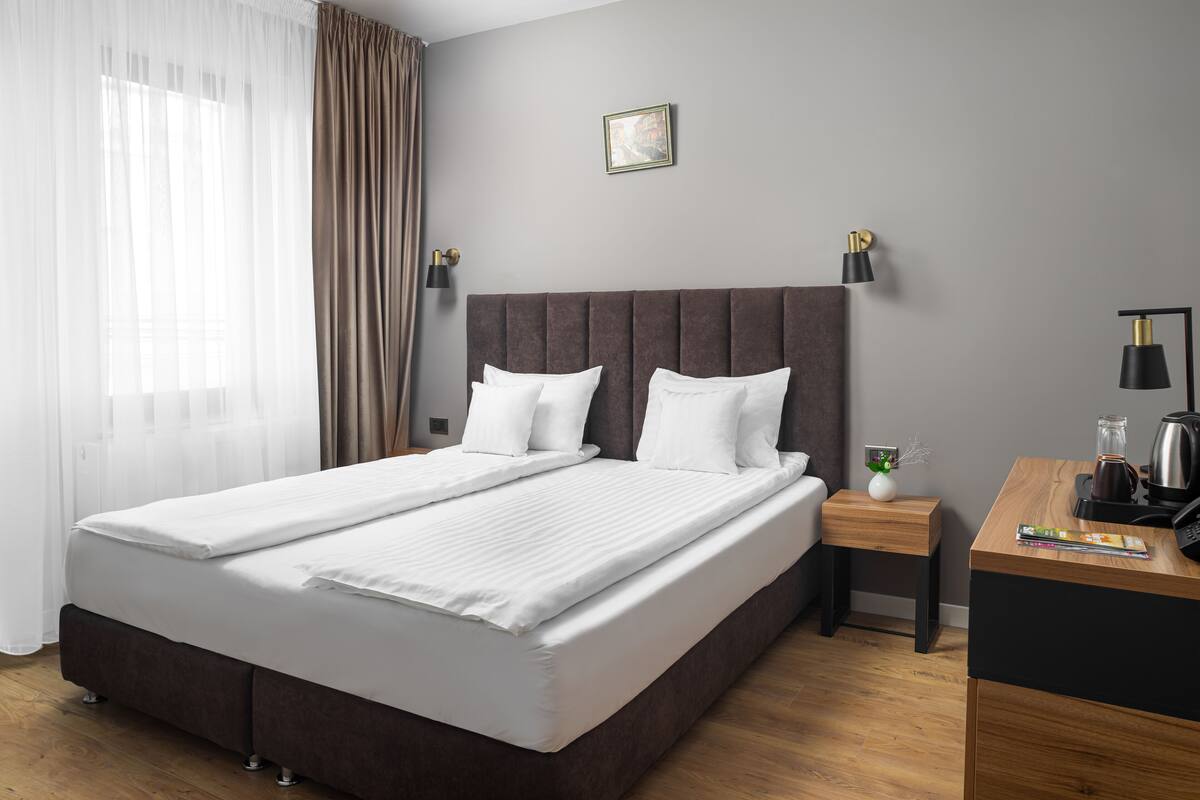 A double bed with white linens, a wooden nightstand with a lamp, and a small table with a coffee pot.