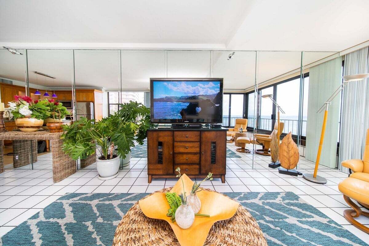 A modern living room with a large flat-screen TV, a dining area with wicker furniture, and a view of the sea through large windows.