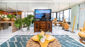 Un soggiorno moderno con una grande televisione a schermo piatto, una zona pranzo con mobili in vimini e vista sul mare attraverso ampie finestre.