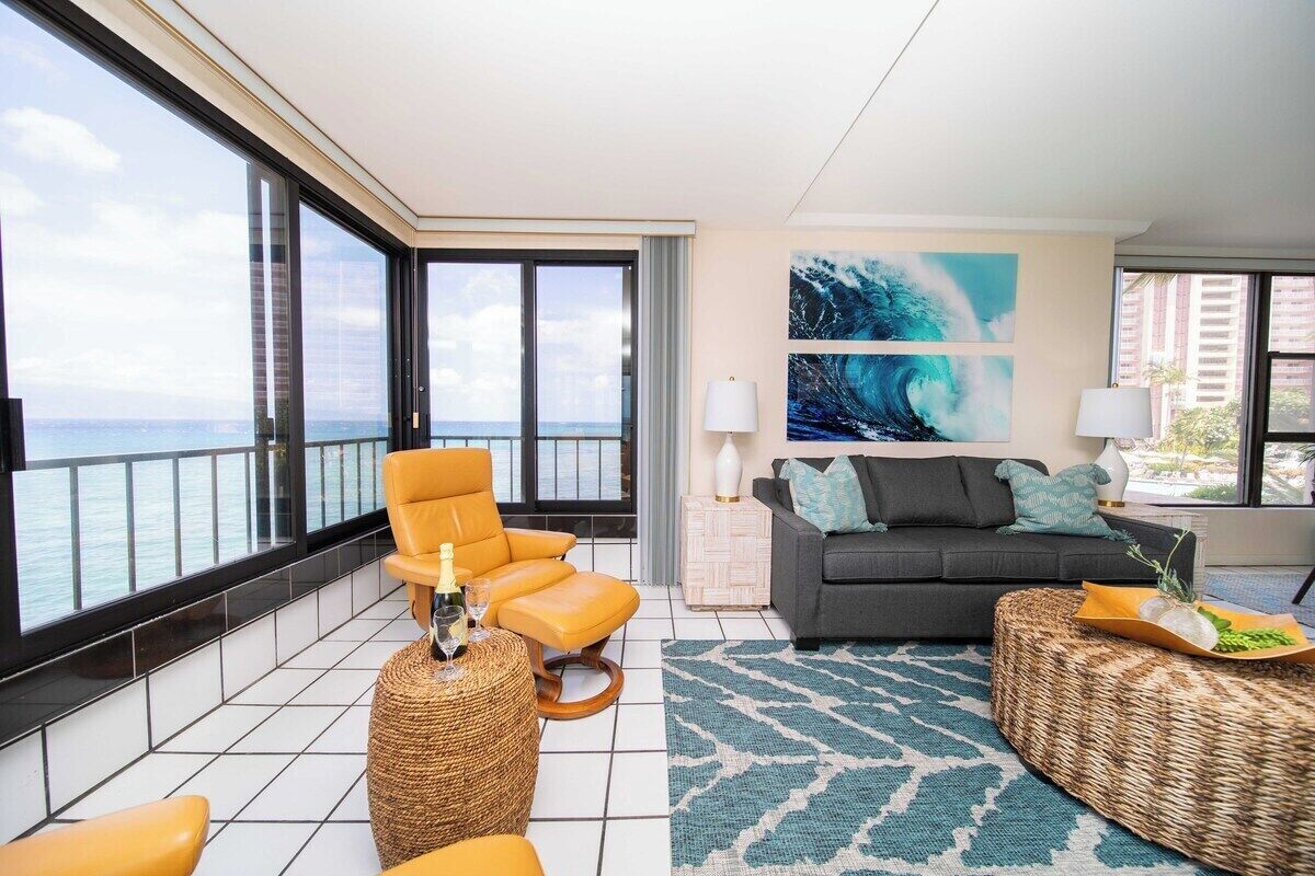 A spacious living room with a balcony overlooking the sea, featuring a sofa, armchair, and a large abstract painting.