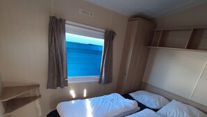 A compact bedroom with a single bed, a window with curtains, and a small shelf unit. - Beautiful mobile home close to the beach and the sea. (IJmuiden)