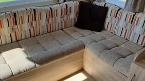 A modern RV interior with a sectional sofa, wooden cabinets, and a large window offering a view of the outdoors. - Beautiful mobile home close to the beach and the sea. (IJmuiden)