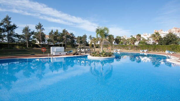 Una piscina amplia y cristalina con sillones de descanso y un pequeño puente.