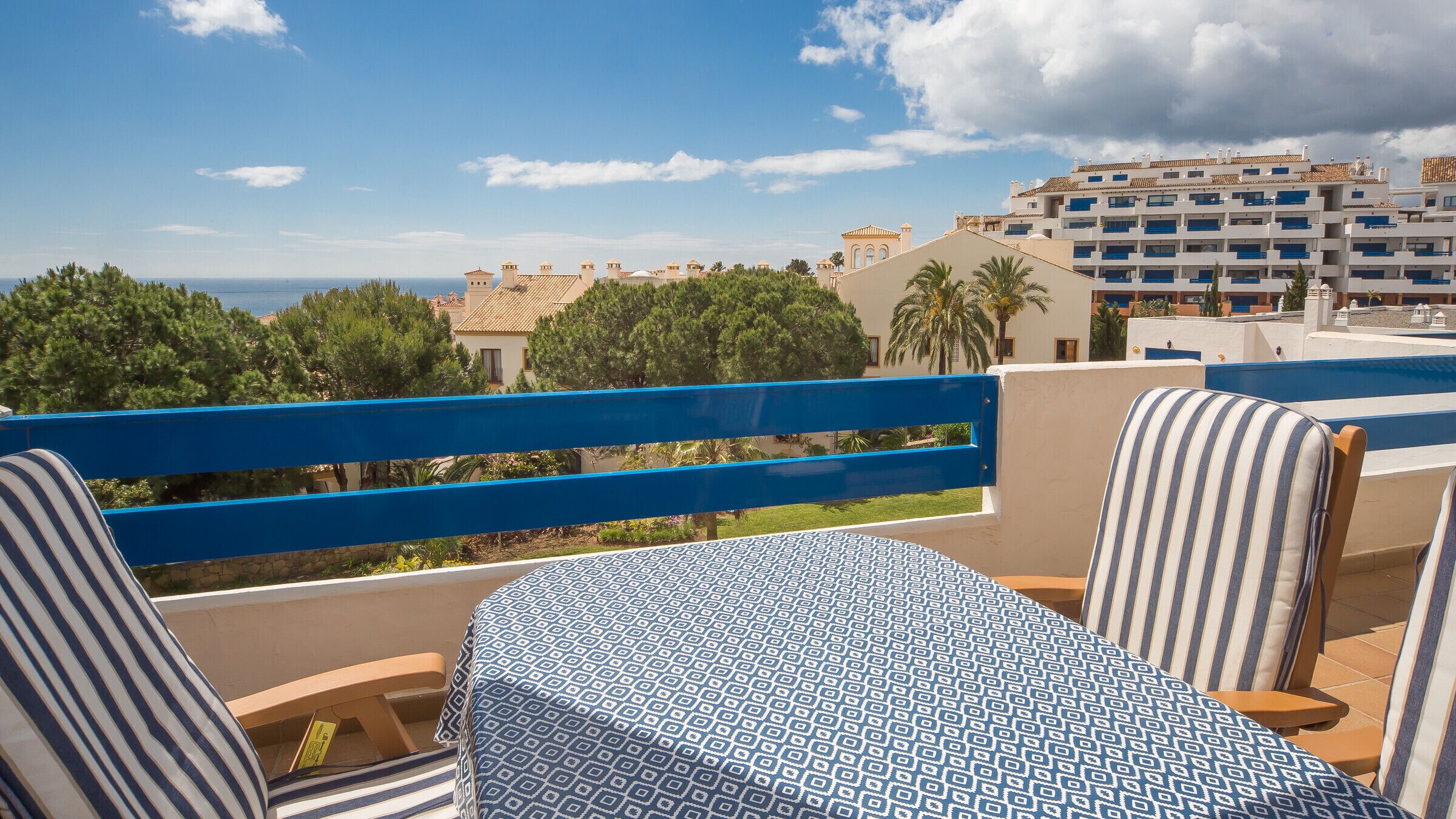 Une terrasse avec des chaises à rayures et une nappe à motifs, donnant sur une zone côtière avec des bâtiments et des arbres.