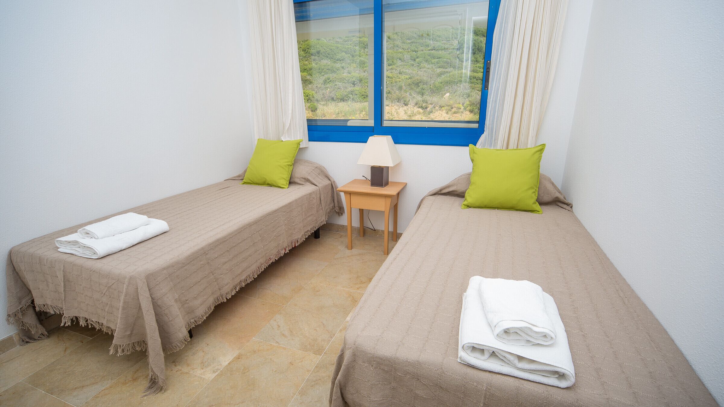 Una habitación con dos camas individuales, cada una con una colcha color beige y un almoháque verde. Entre las camas hay una mesita de noche de madera con una lámpara.