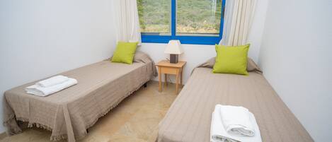 Una habitación con dos camas individuales, cada una con una colcha color beige y un almoháque verde. Entre las camas hay una mesita de noche de madera con una lámpara.