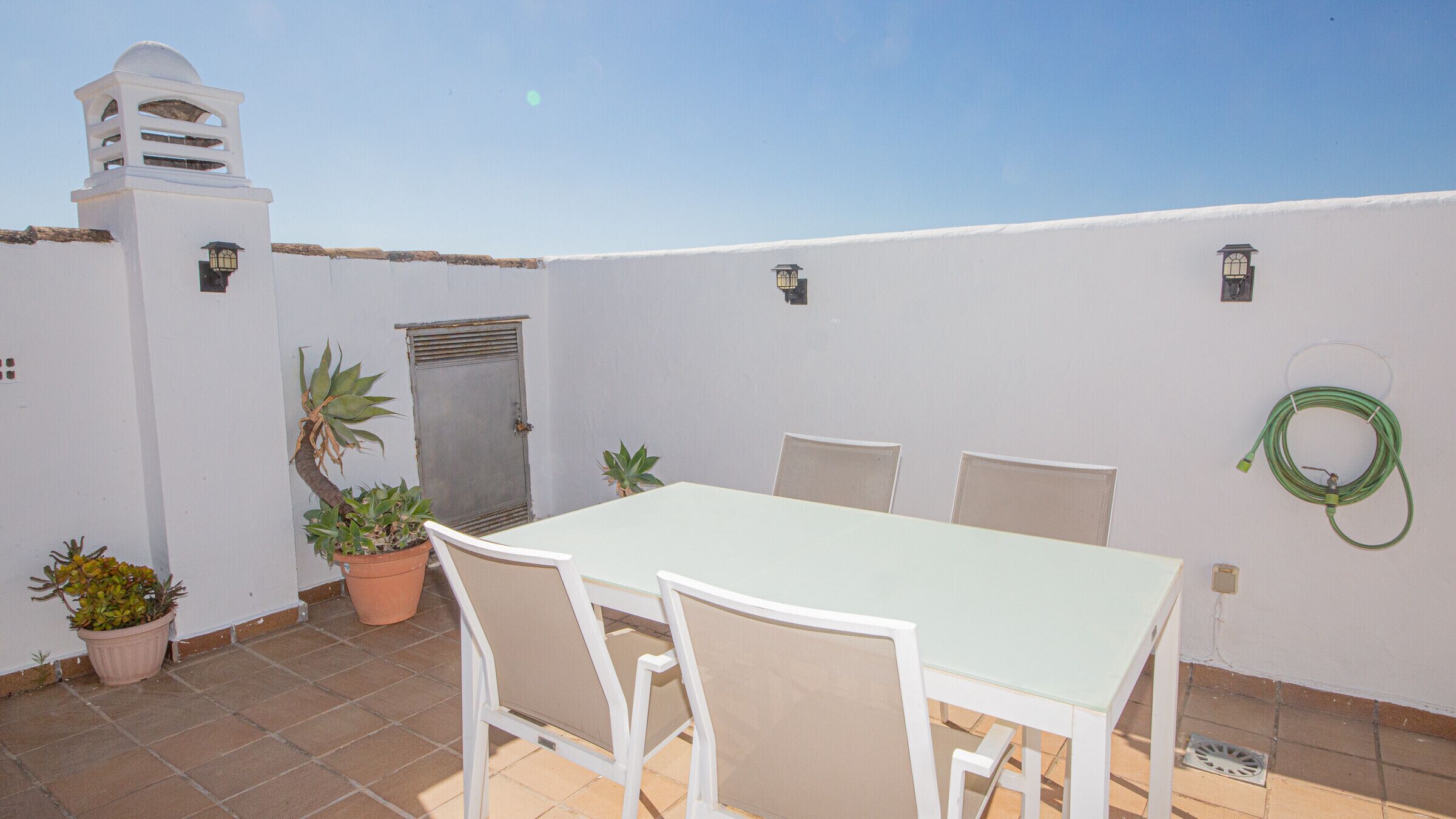 A patio with a table and chairs, potted plants, and a hose on the wall.