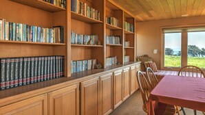 A library with wooden bookshelves filled with books, a dining area with a table and chairs, and a large window offering a view of the outdoors. - Wild Moor - Hot Tub & Star View Retreat (Sea Ranch)