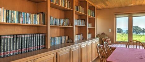 Una biblioteca con estanterías de madera llenas de libros, un comedor con una mesa y sillas, y un ventanal que ofrece vistas al exterior.