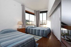 A bedroom with two beds, a nightstand, a lamp, and large windows.