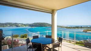 Ein Balkon mit rundem Tisch und Stühlen, der auf eine türkisfarbene Bucht mit klarem Wasser und einer entfernten Küste blickt.