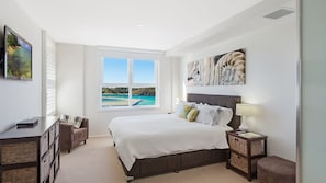 A bedroom with a large bed, a desk, and a chair, a window with a view of the beach, and artwork on the wall. - Wharf Apartment Unit 7 (NAROOMA)