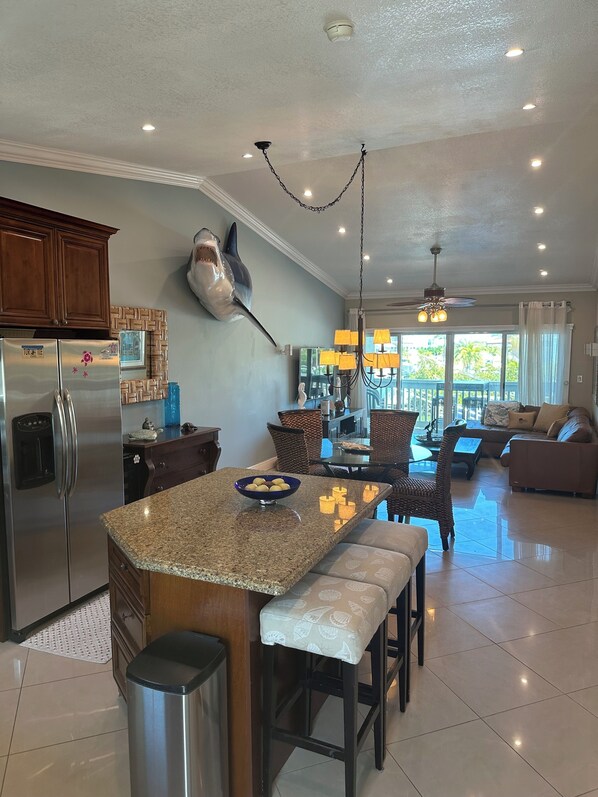 A modern kitchen with a built-in refrigerator, a large island with a bowl of fruit, and a dining area with wicker furniture. - Key Largo Paradise - Beautiful Key Largo Oceanside Townhouse - 28 Day Min Rent (Key Largo)
