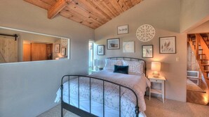 A bedroom with a bed, bedside tables, a mirror, artwork on the walls, and a wooden ceiling.