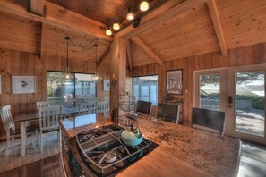 A wooden interior with a kitchen area, dining table, chairs, and a large window providing a view of the outdoors. - Point of View | Dog-Friendly Oceanfront Escape (Sea Ranch)