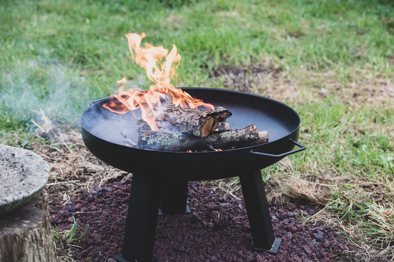 Una parrilla portátil negra con llamas visibles y leña ardiendo, sobre una superficie cubierta de césped.