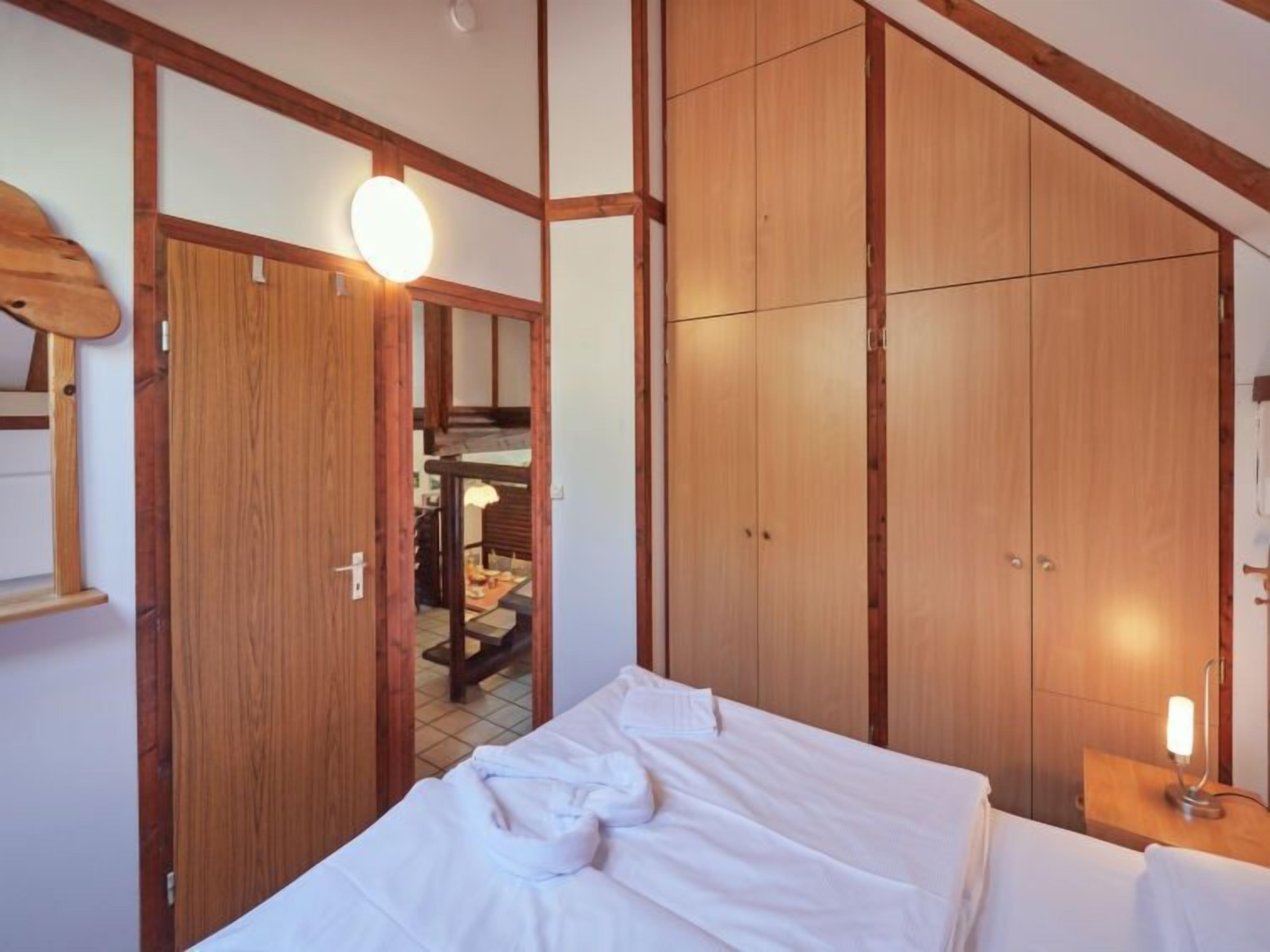 A hotel room with a bed, wooden wardrobe, and a small table with a lamp.