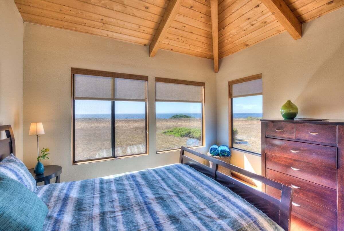 A bedroom with a large bed, wooden ceiling beams, a dresser, and a view of the beach.