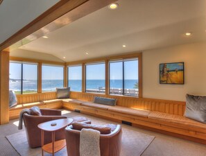 A room with a large window overlooking the sea, a wooden bench seating area, and a painting on the wall.