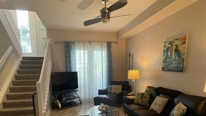 A living room with a sofa, two armchairs, a coffee table, a TV, a ceiling fan, a painting, and a staircase.