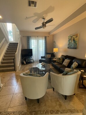 A living room with a sofa, two armchairs, a coffee table, a TV, a ceiling fan, a painting, and a staircase.