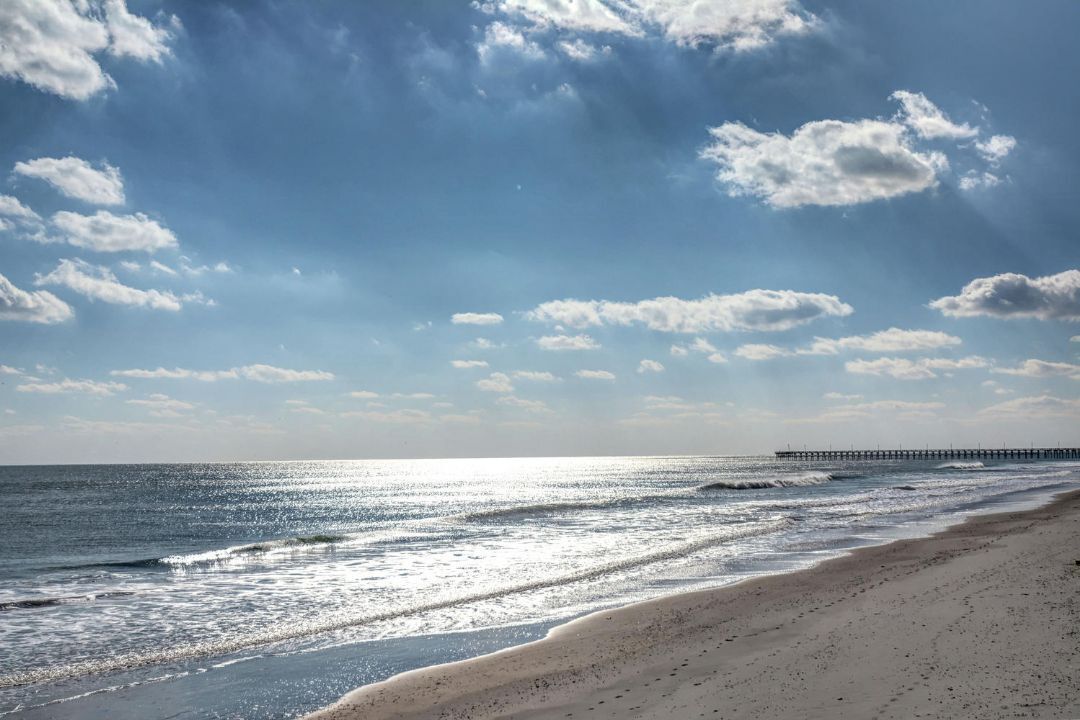 Une plage avec une jetée qui s’avance dans l’océan, un ciel dégagé et la lumière du soleil qui se reflète sur l’eau.