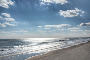 Uma praia com um píer que se estende para o oceano, céu claro e reflexos do sol na água.