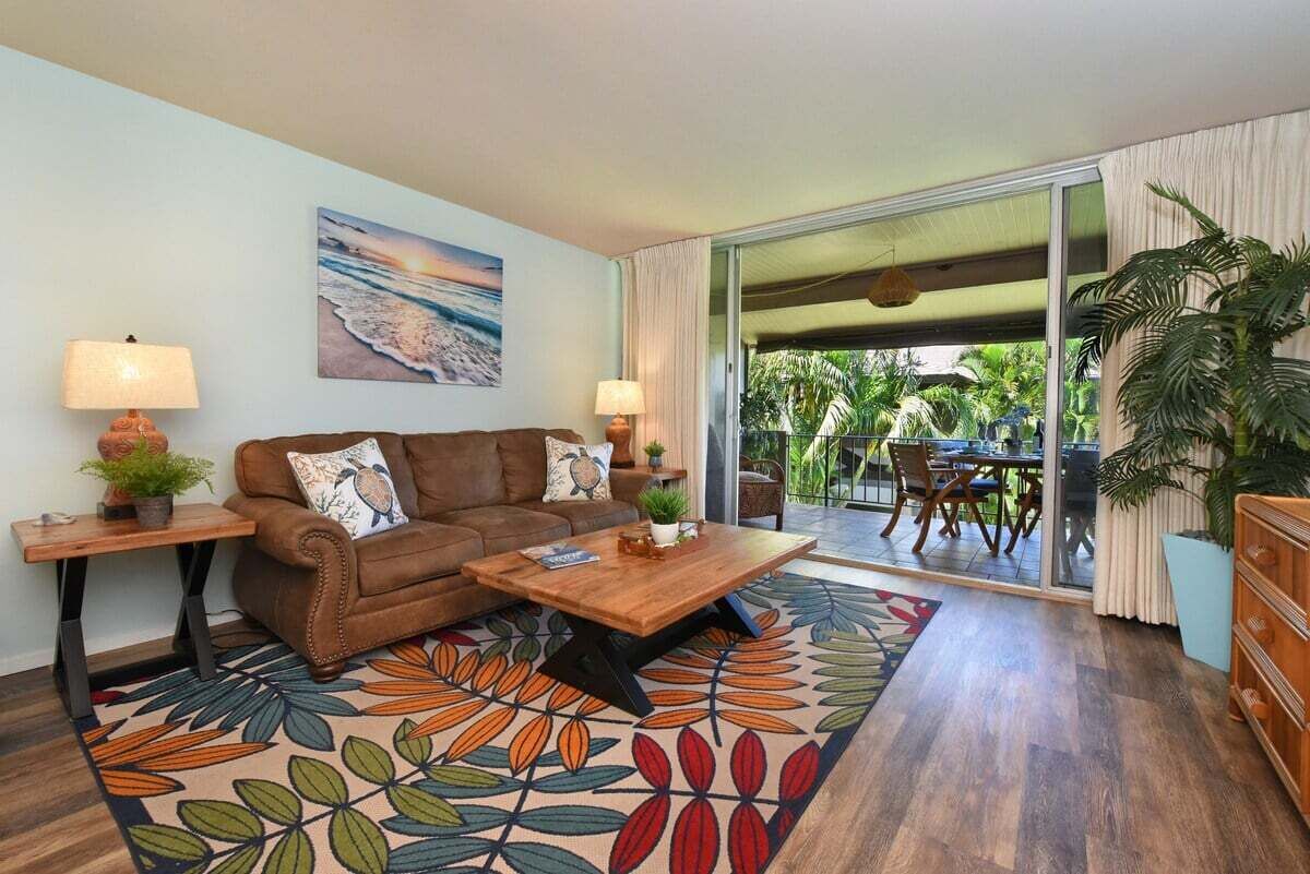 Una sala de estar con un sofá marrón, dos mesas auxiliares con lámparas, una mesa de centro y un tapete colorido. Hay un cuadro en la pared, una puerta corrediza de vidrio que da a un área exterior con una mesa y sillas.