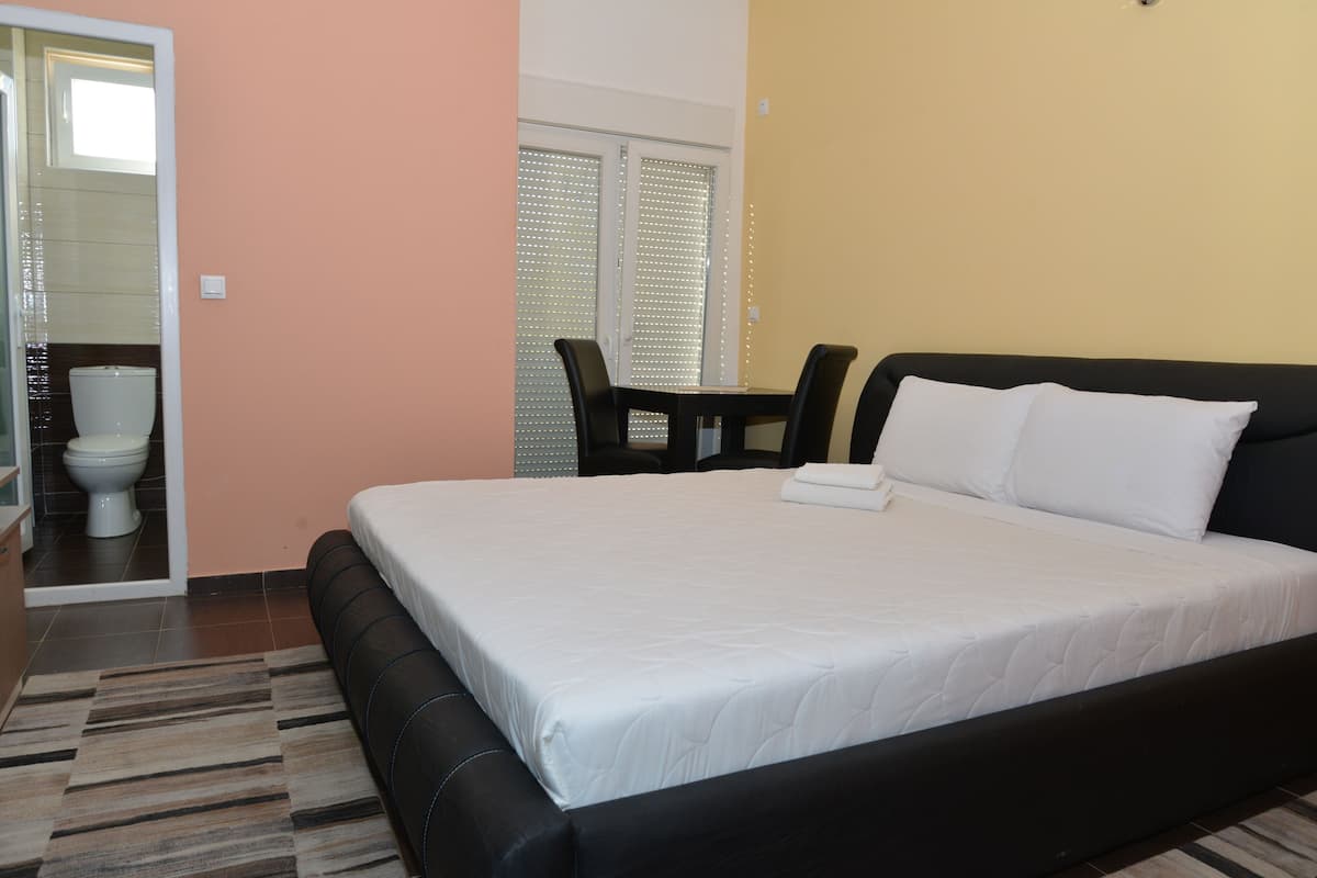 Ein Schlafzimmer mit einem großen Bett, einem kleinen Tisch und einem Stuhl. Ein Badezimmer ist durch eine geöffnete Tür sichtbar.