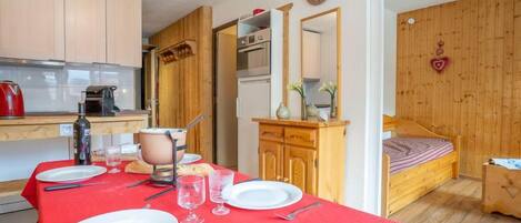 Una habitación acogedora con una mesa de comedor para dos, una cama con cabecera de madera y una kitchenette con armarios de madera y un hervidor rojo.