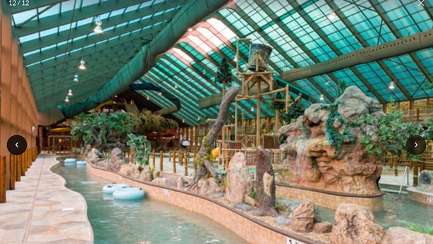 Parque acuático interior con techo de cristal, formaciones rocosas artificiales y flotadores inflables.