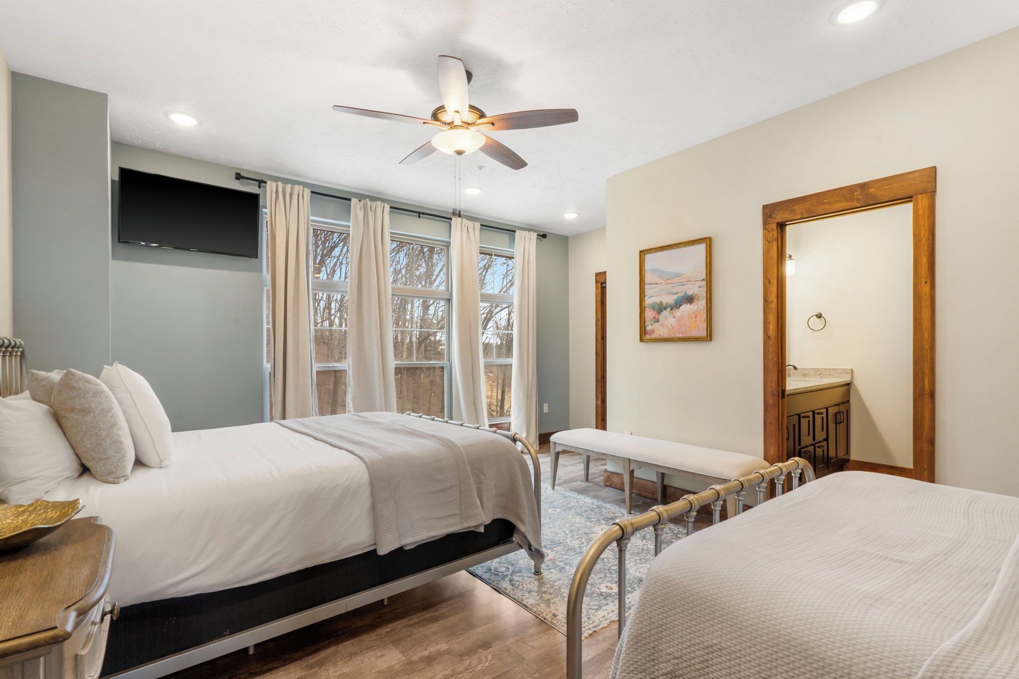 A bedroom with two beds, a ceiling fan, a TV, a window with curtains, a painting, and a wooden-framed mirror.