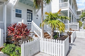 A white multi-story building with a white picket fence, a staircase, and various plants.