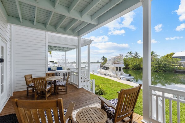 A covered outdoor deck with wooden furniture, a view of a marina, and a grassy area.