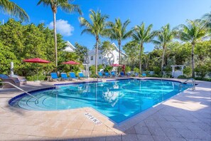 A swimming pool with lounge chairs and umbrellas, surrounded by palm trees and a white building.