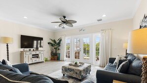 Una sala de estar con ventilador de techo, televisor, sofá, mesa de centro y vistas a través de puertas de cristal.