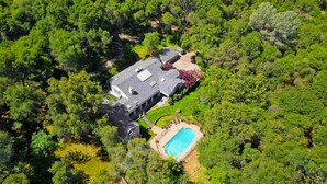An aerial view of a house surrounded by dense greenery, with a swimming pool and a covered patio area.