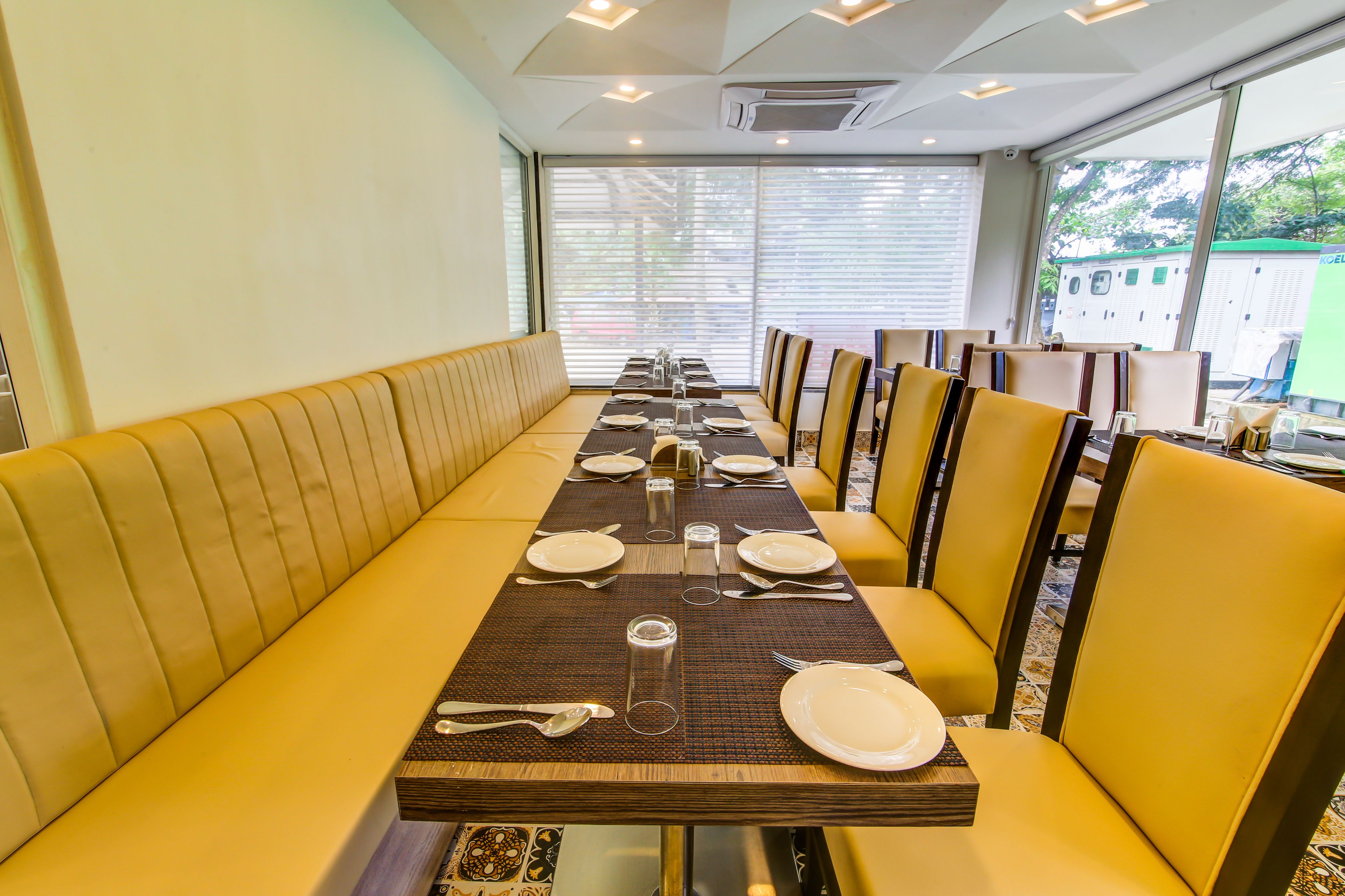 Una larga mesa de comedor con bancos acolchados amarillos, dispuesta con platos, cubiertos y vasos.