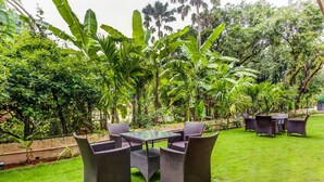 Un patio con muebles de mimbre y una mesa de vidrio rodeado de exuberante vegetación y altas plantas de banano.
