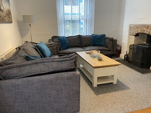 Een woonkamer met een zithoek, een salontafel, een open haard en een raam met jaloezieën.