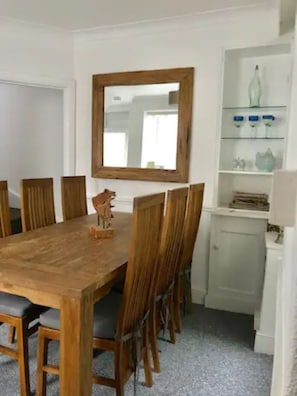 Een eetruimte met een houten tafel en stoelen, een spiegel boven de tafel, en een witte kast met planken.