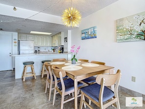 A modern kitchen with white cabinets, a central island, and a dining area with wooden chairs and blue cushions.