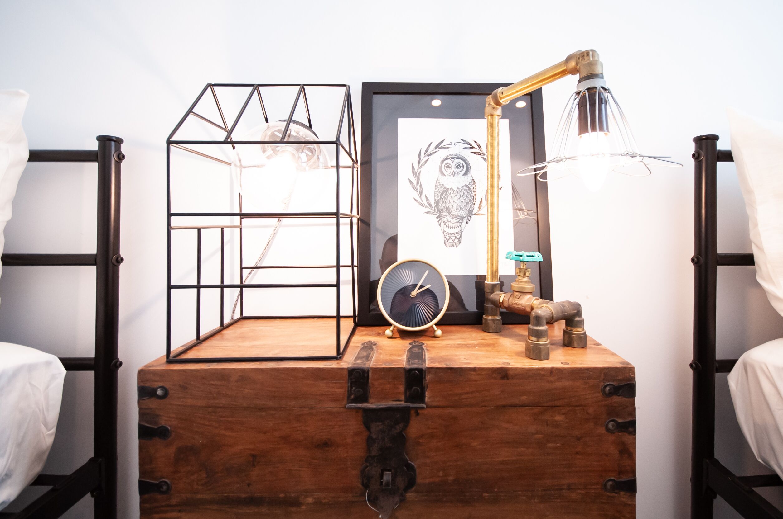 A wooden chest with a clock and a lamp on top, a framed picture of an owl, and a metal rack on a bed.