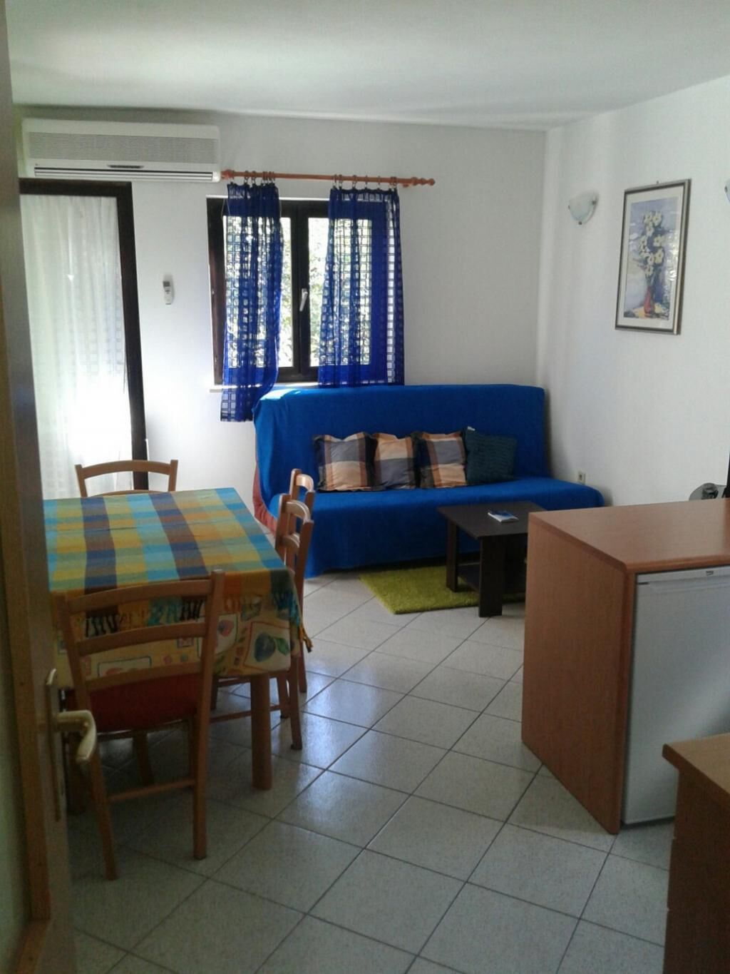 A compact living space with a blue sofa, a dining table with chairs, a small kitchenette, and a window with blue curtains.